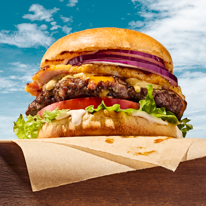 Closeup image of a beef burger with red onion, peameal bacon, cheese, tomato, lettuce and dijon mustard between two slices of brioche burger buns.