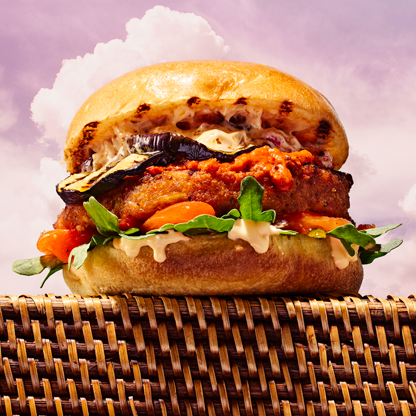 Closeup image of a chickpea veggie burger with eggplant, baba ganouj, burger sauce, muffuletta olive antipasto, arugula and roasted red peppers between two slices of brioche burger buns.