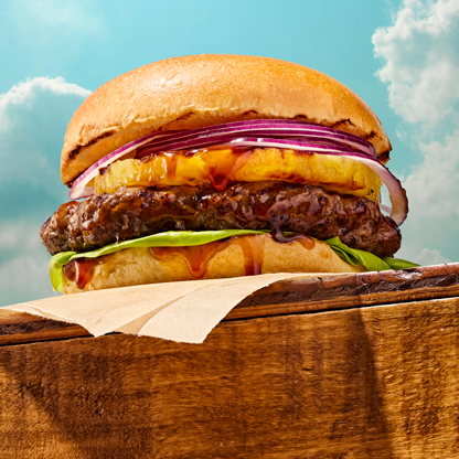 Closeup image of a beef burger with red onion, pineapple, lettuce, teriyaki grilling sauce between two slices of brioche burger buns.